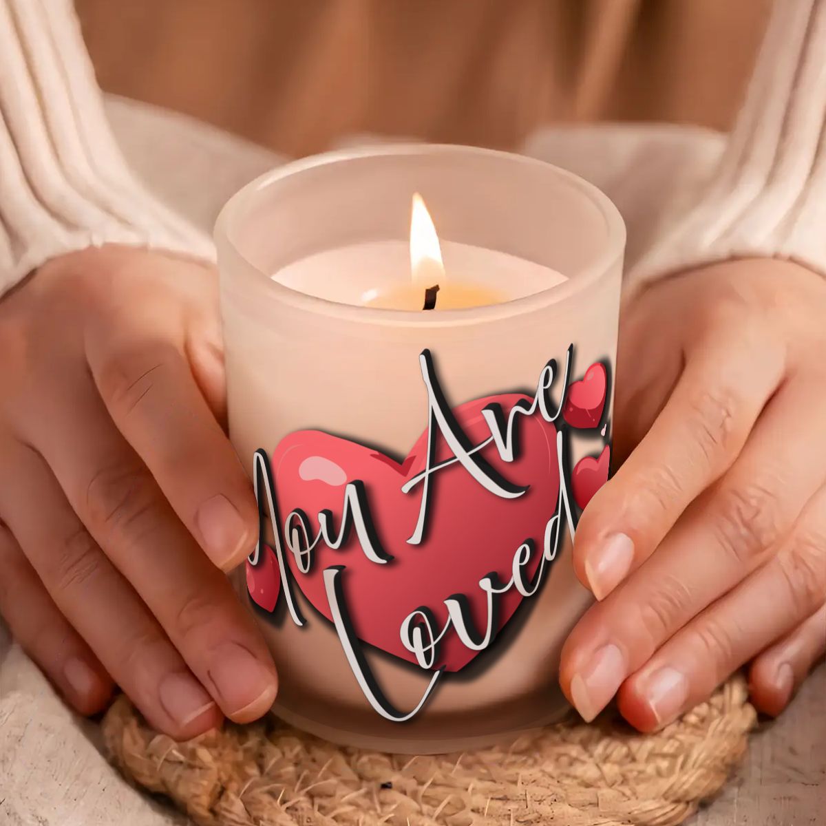 Candle with 'You Are Loved' text held by hands on a woven mat