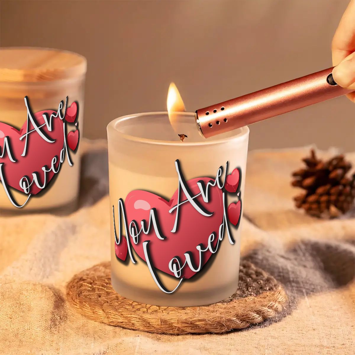 Candle being lit with 'You Are Loved' text on a heart design, surrounded by pinecones.