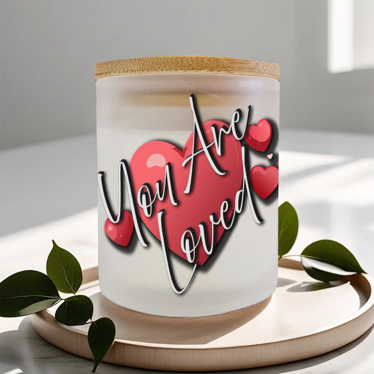 Candle with 'You Are Loved' text and heart design on a white surface with green leaves.